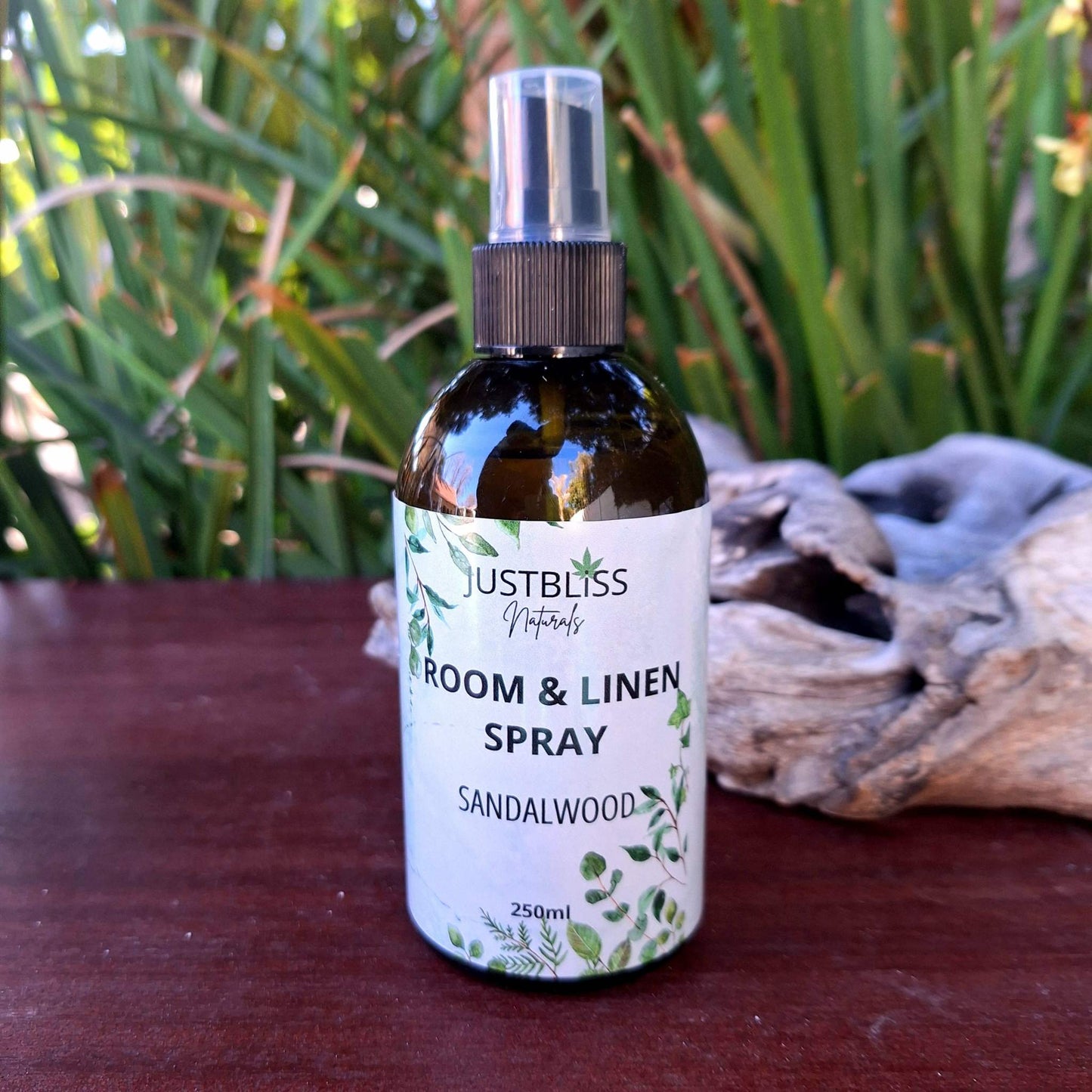 ROOM & LINEN SPRAY bottle with sandalwood scent placed on a wooden surface with greenery background.