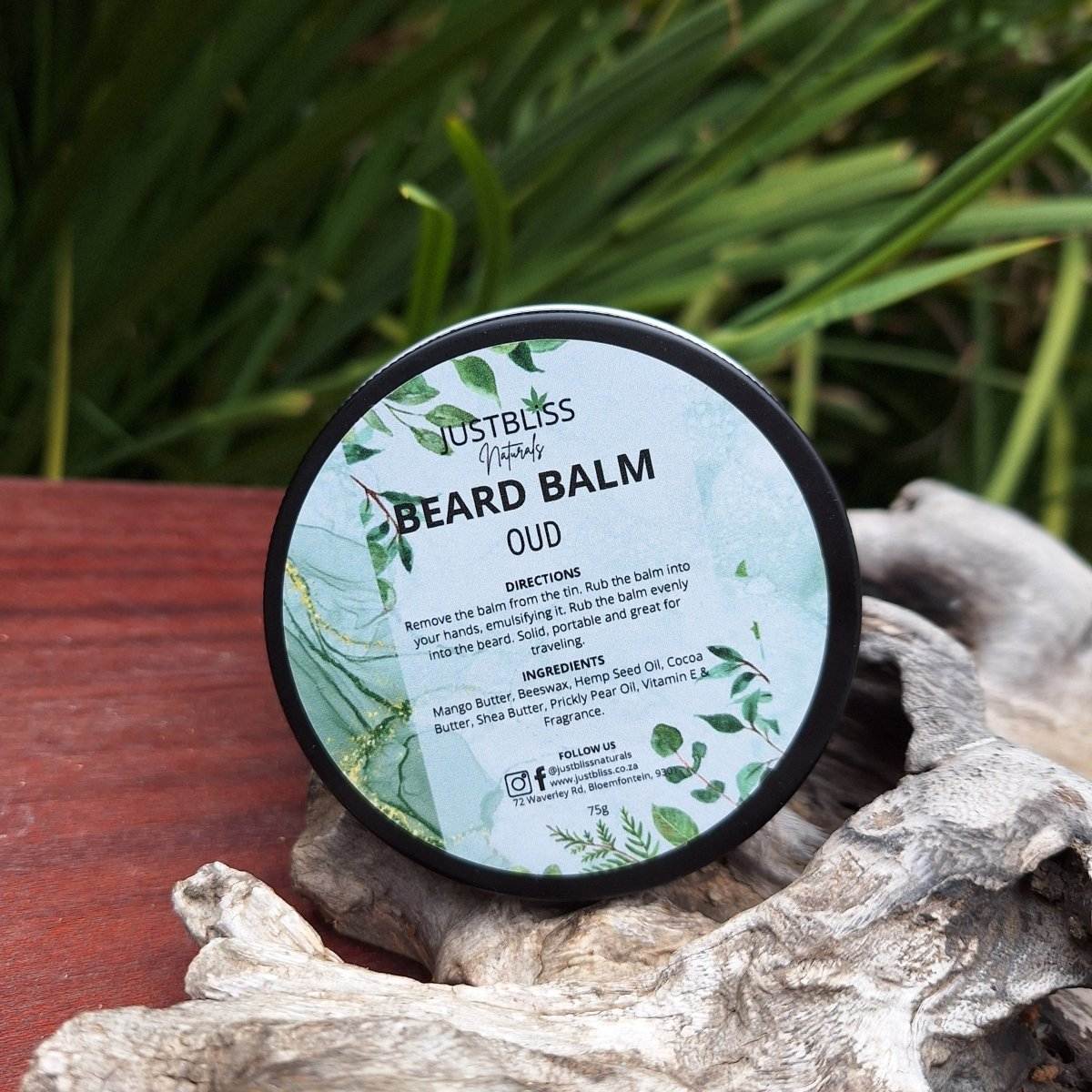 JustBliss Oud Beard Balm tin on a wooden surface with green foliage background.