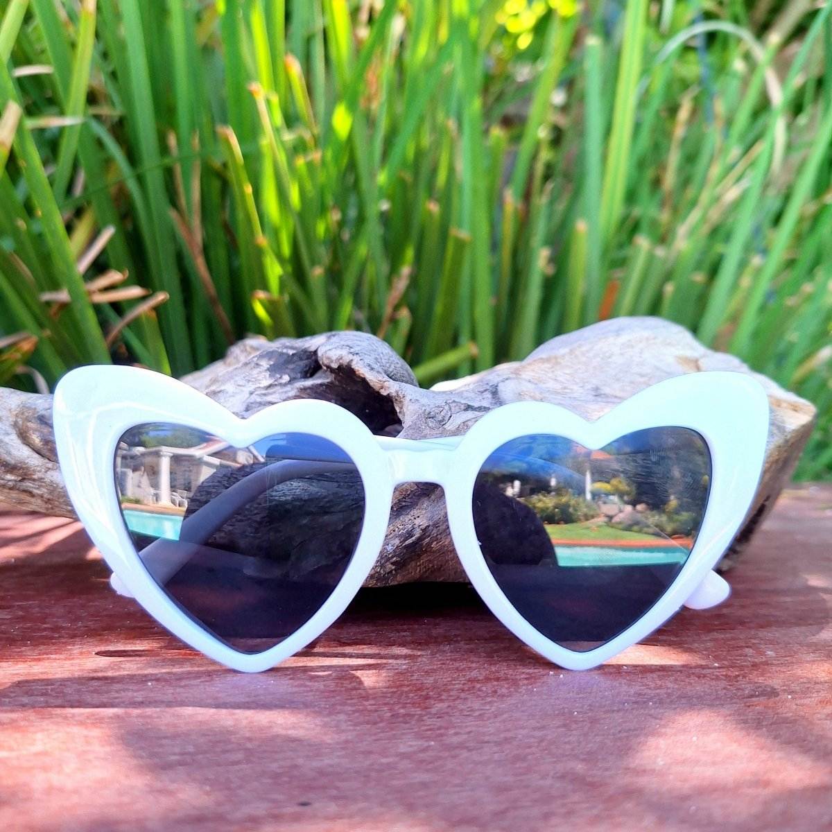 Heart-shaped sunglasses from Bride To Be Gift Box 1 on wooden surface with greenery backdrop.