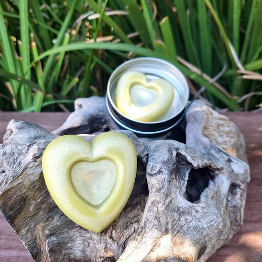 Heart-shaped lotion bar in a reusable tin, set on rustic wood with greenery in the background.